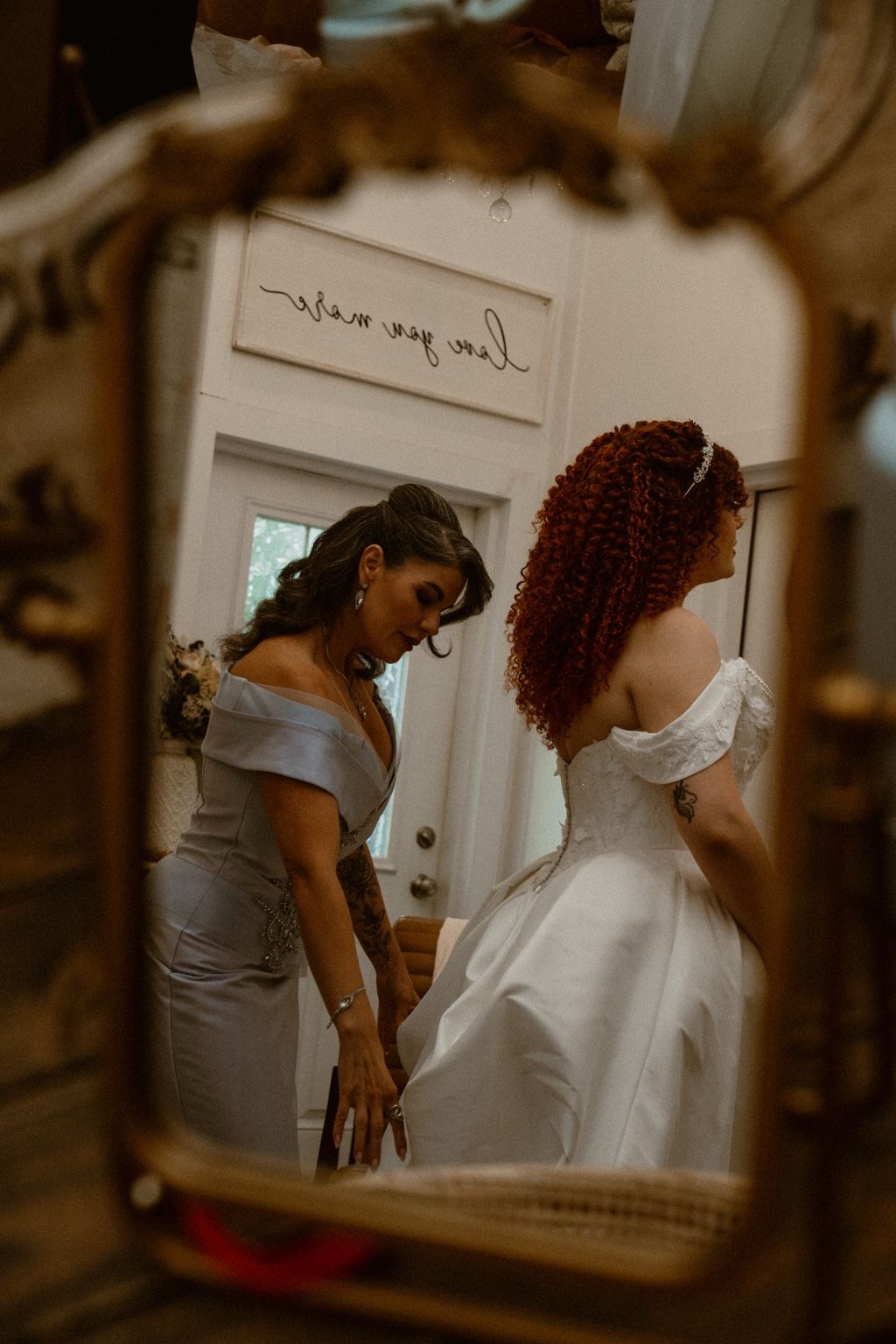 A bride being helped with her gown, reflected in an antique mirror