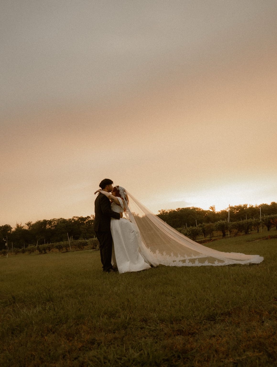 A couple embracing in an open field at sunset, the bride's long veil trailing across the grass
